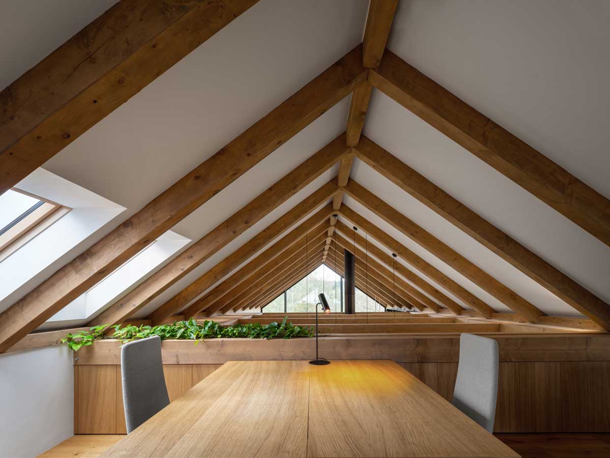 Pracovna v podkroví moderní chaty s masivními dřevěnými trámy a stolem pod šikmou střechou