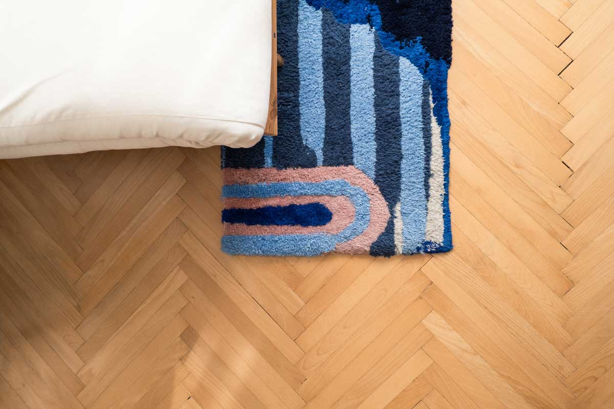 Barevný ručně tkaný koberec položený na dřevěných parketách vedle postele.