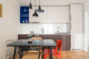 Moderní kuchyň s výraznou modrou policí, kombinací světlých a tmavých dvířek a jídelním stolem se směsí designových židlí.