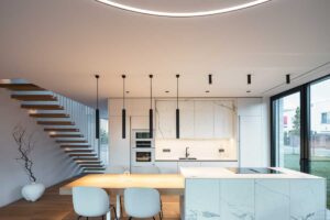 Moderní kuchyň s mramorovými obklady, dřevěnou podlahou a velkoformátovým oknem, které vizuálně propojuje interiér s okolní zahradou.