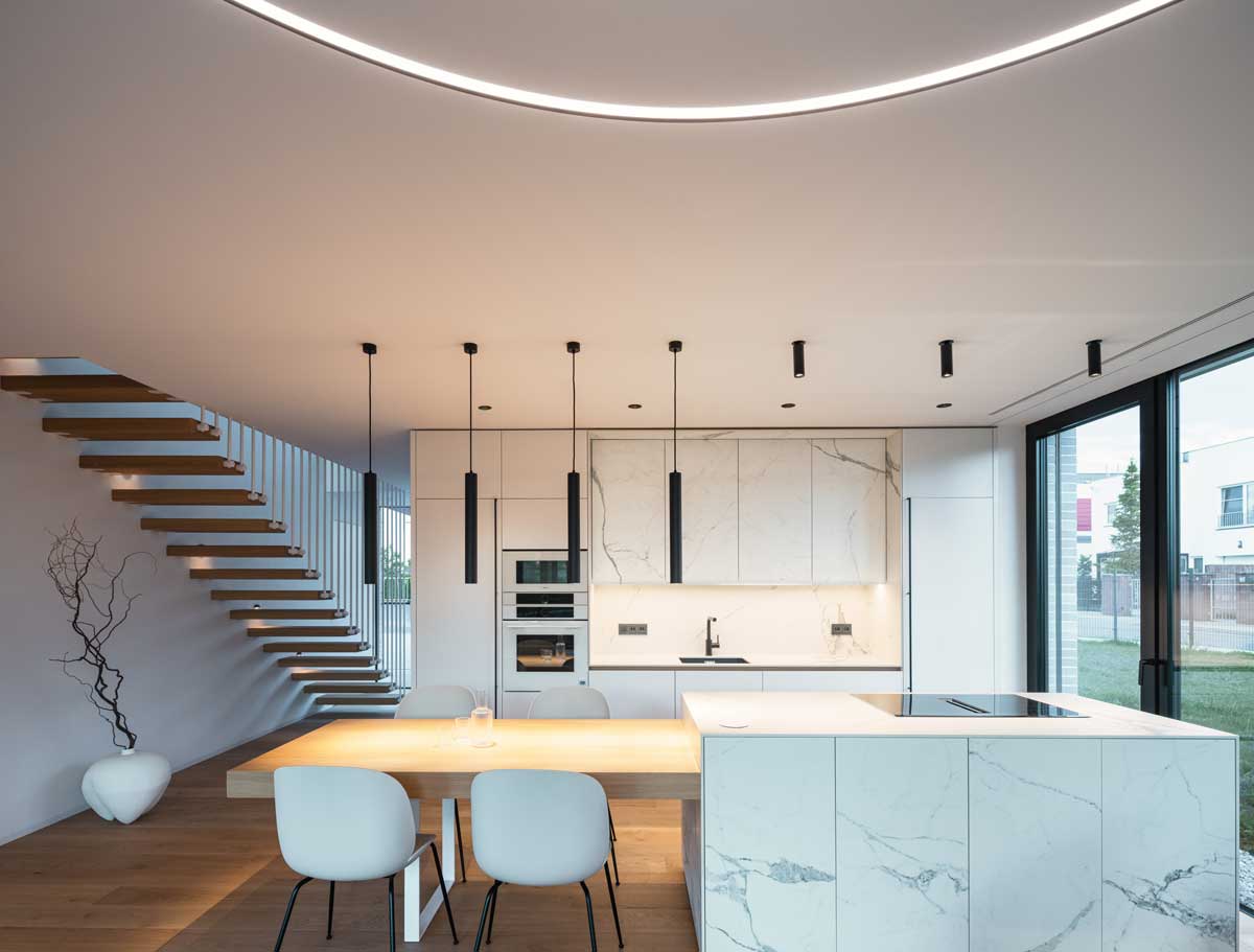 Moderní kuchyň s mramorovými obklady, dřevěnou podlahou a velkoformátovým oknem, které vizuálně propojuje interiér s okolní zahradou.