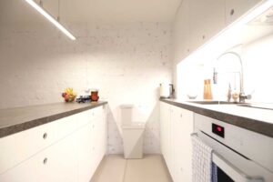 Světlá garsonková kuchyně s bílými skříňkami, hlubokými zásuvkami a tmavou pracovní deskou.