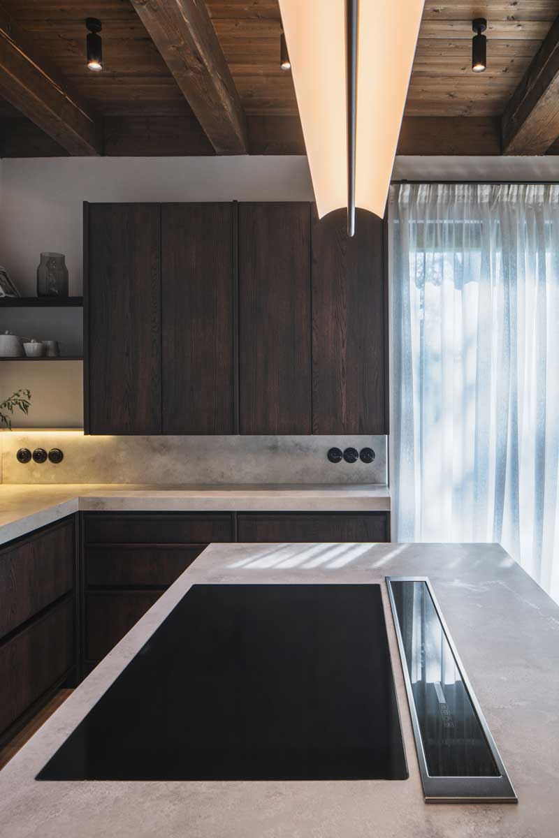 Kuchyně s tmavým dřevem, kamennou deskou a trámovým stropem v moderní horské chalupě