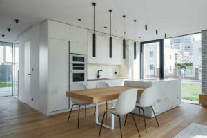 Propojená kuchyň a jídelna s dubovým stolem navazujícím na kuchyňský ostrůvek a velkými okny přivádějícími denní světlo.