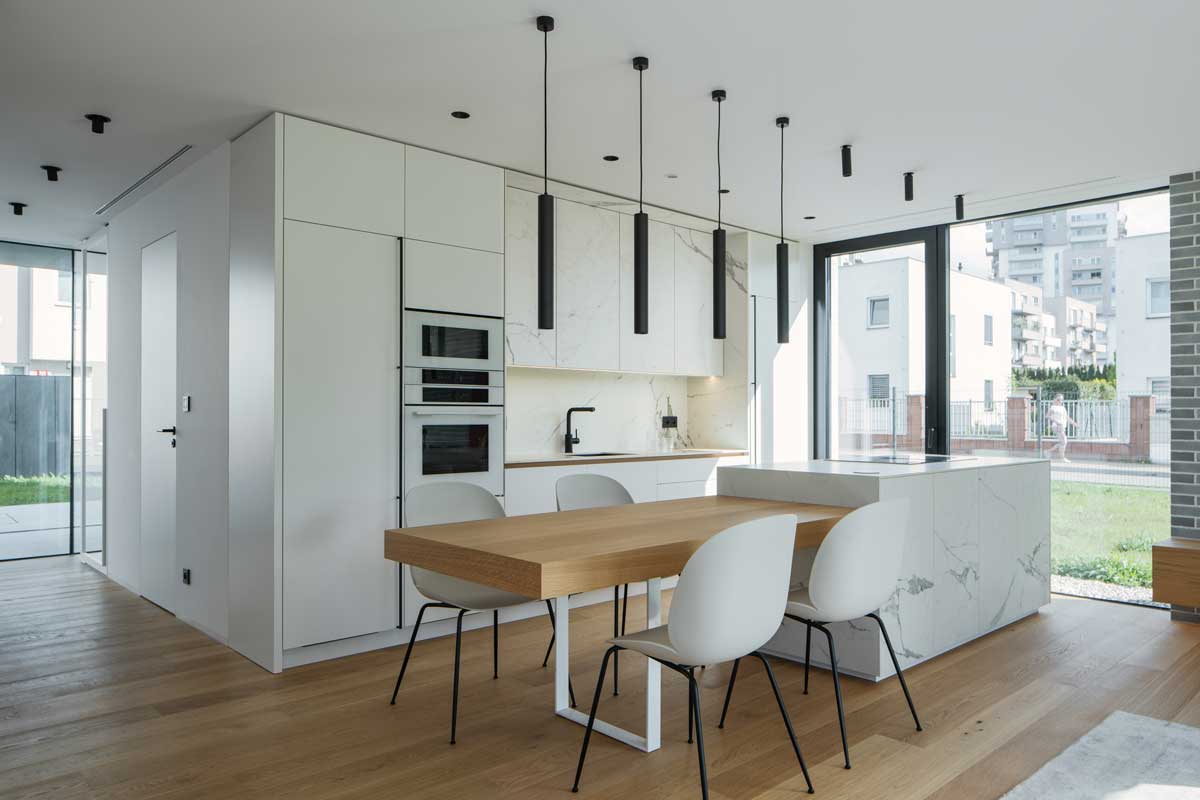 Propojená kuchyň a jídelna s dubovým stolem navazujícím na kuchyňský ostrůvek a velkými okny přivádějícími denní světlo.