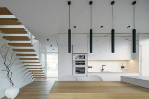Minimalistická kuchyň s bílými a mramorovými plochami, černými svítidly a otevřeným schodištěm z masivního dřeva.