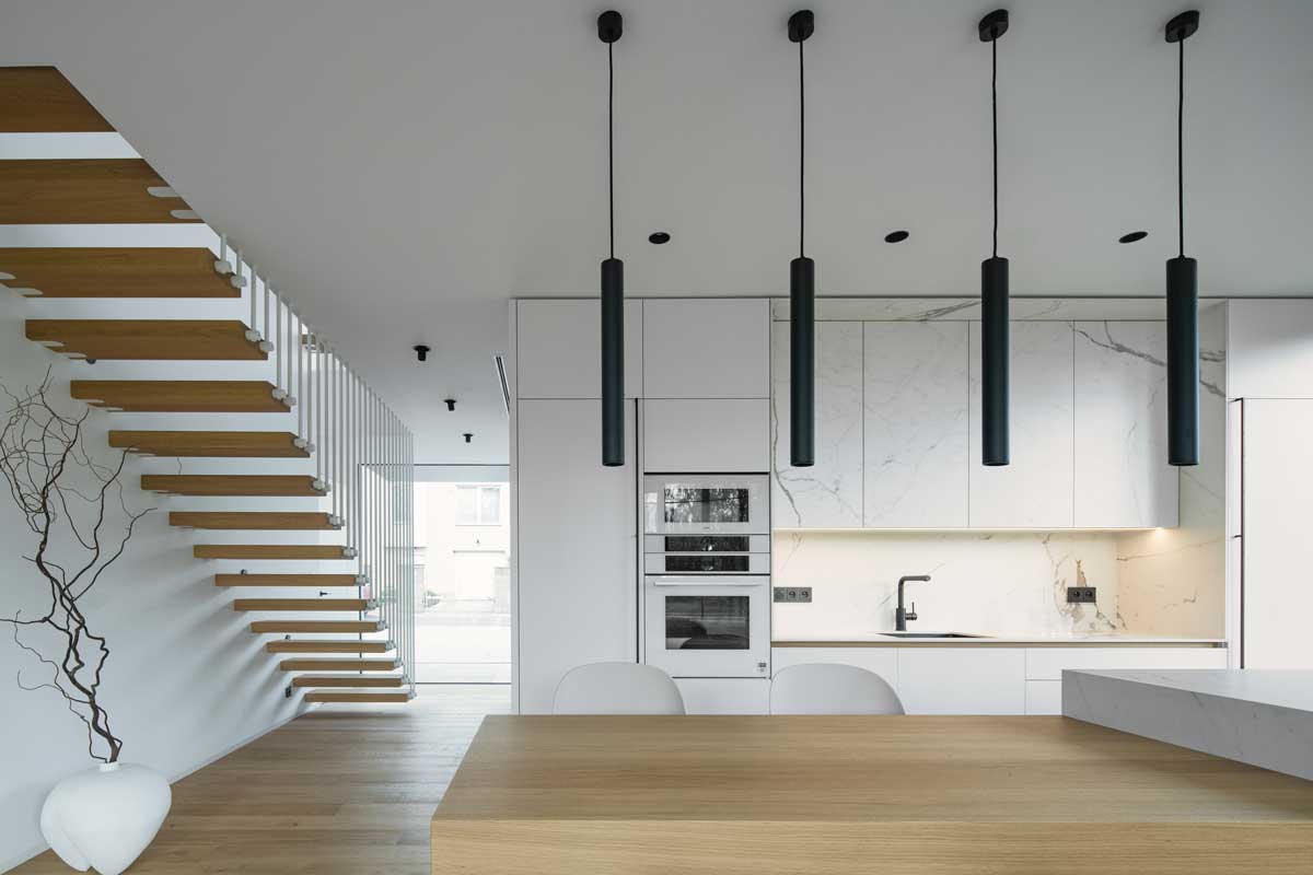 Minimalistická kuchyň s bílými a mramorovými plochami, černými svítidly a otevřeným schodištěm z masivního dřeva.