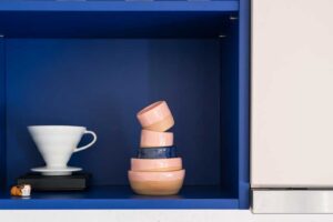 Modrá kuchyňská police s dekorativním sklem, keramickými miskami a minimalistickým kávovým vybavením.