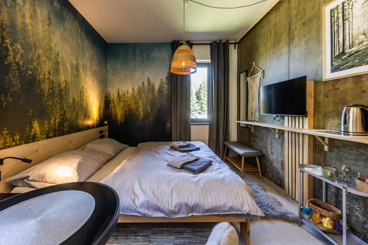Ložnice v horské chatě s fototapetou lesa, betonovou stěnou a dřevěným nábytkem