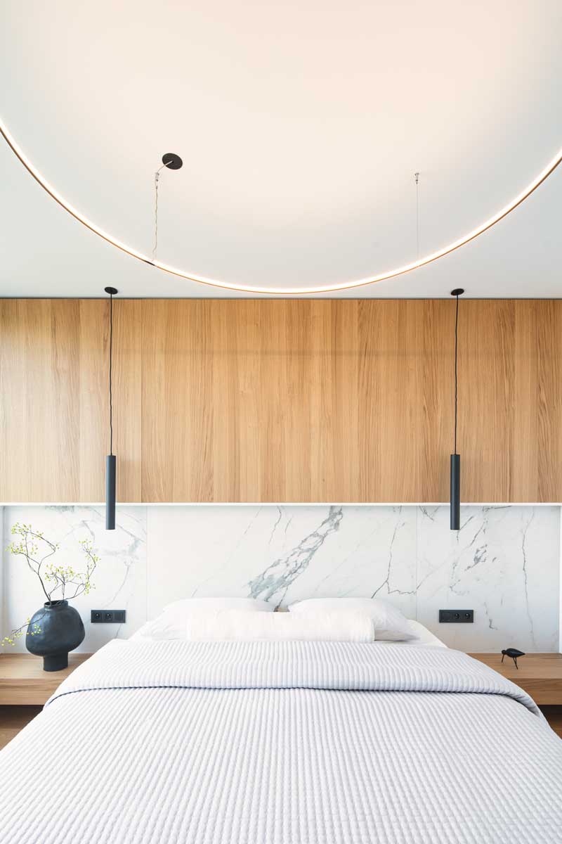 Designová ložnice s mramorovým obkladem za čelem postele, dýhovaným panelem a závěsnými černými svítidly.