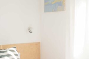 Světlá ložnice s minimalistickou kompozicí, obrazem v modrých tónech a dřevěnou postelí.