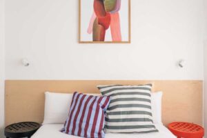 Kompaktní ložnice s dřevěným čelem postele, pruhovanými polštáři a výrazným abstraktním obrazem.