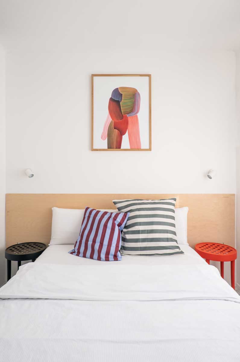 Kompaktní ložnice s dřevěným čelem postele, pruhovanými polštáři a výrazným abstraktním obrazem.