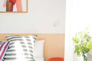 Ložnice s proužky světla, barevnými polštáři a rostlinou na parapetu.