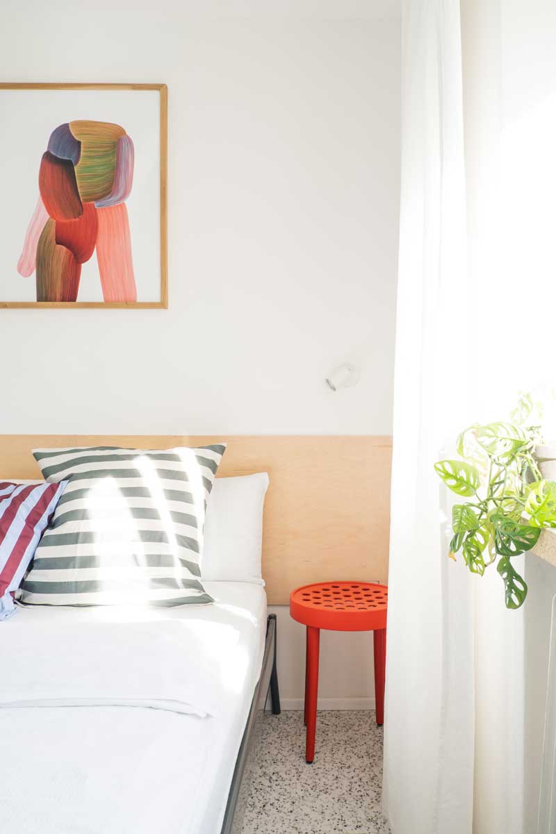 Ložnice s proužky světla, barevnými polštáři a rostlinou na parapetu.