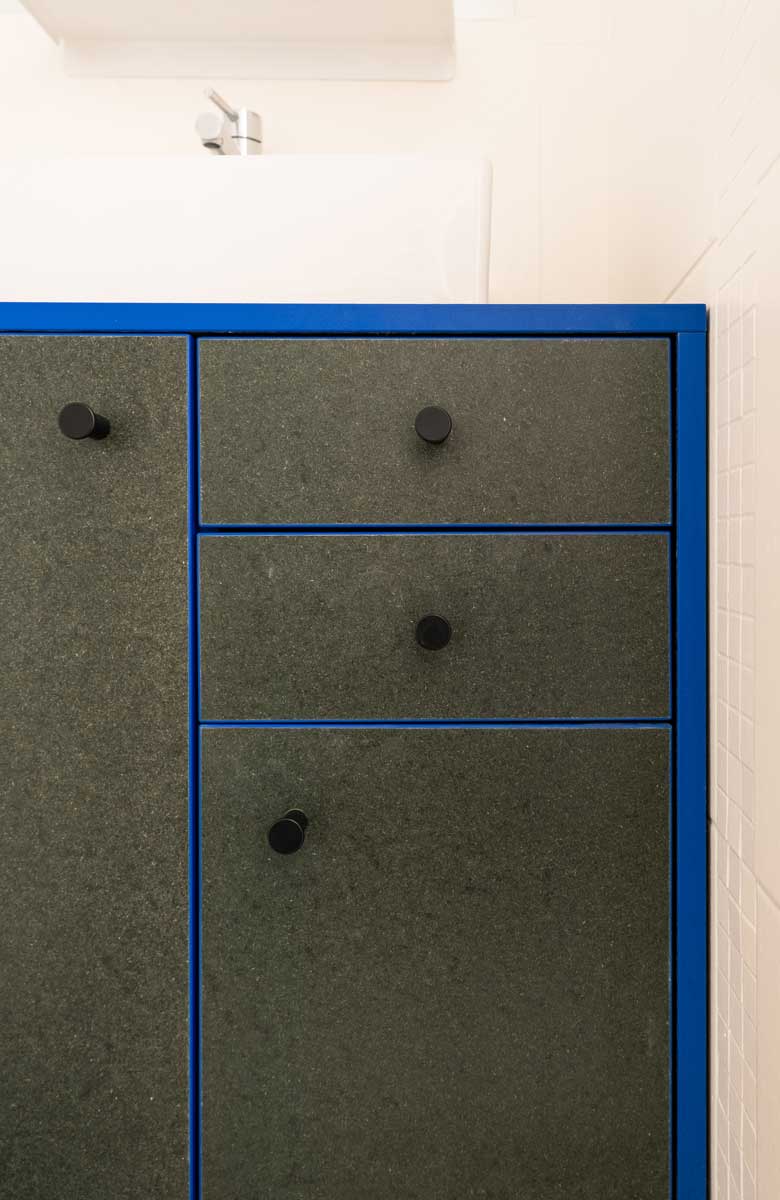 Detail koupelnové skříňky s modrým rámem a tmavými dvířky z recyklovaného materiálu.