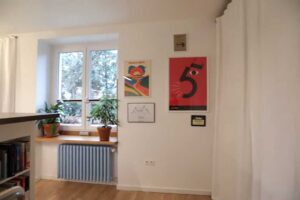 Stěna v obývací části garsonky s barevnými plakáty a pokojovou rostlinou u okna.