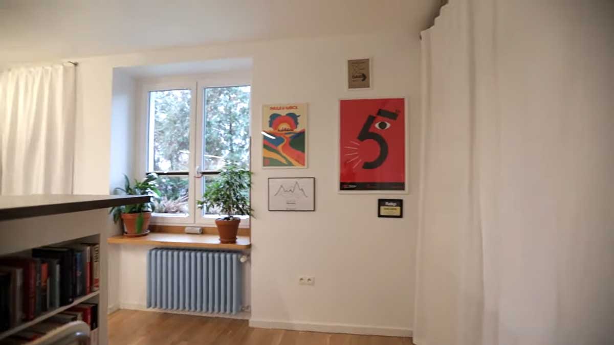 Stěna v obývací části garsonky s barevnými plakáty a pokojovou rostlinou u okna.