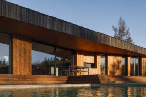 Pohled na dřevěnou fasádu domu od bazénu s krytou terasou a velkými okny.