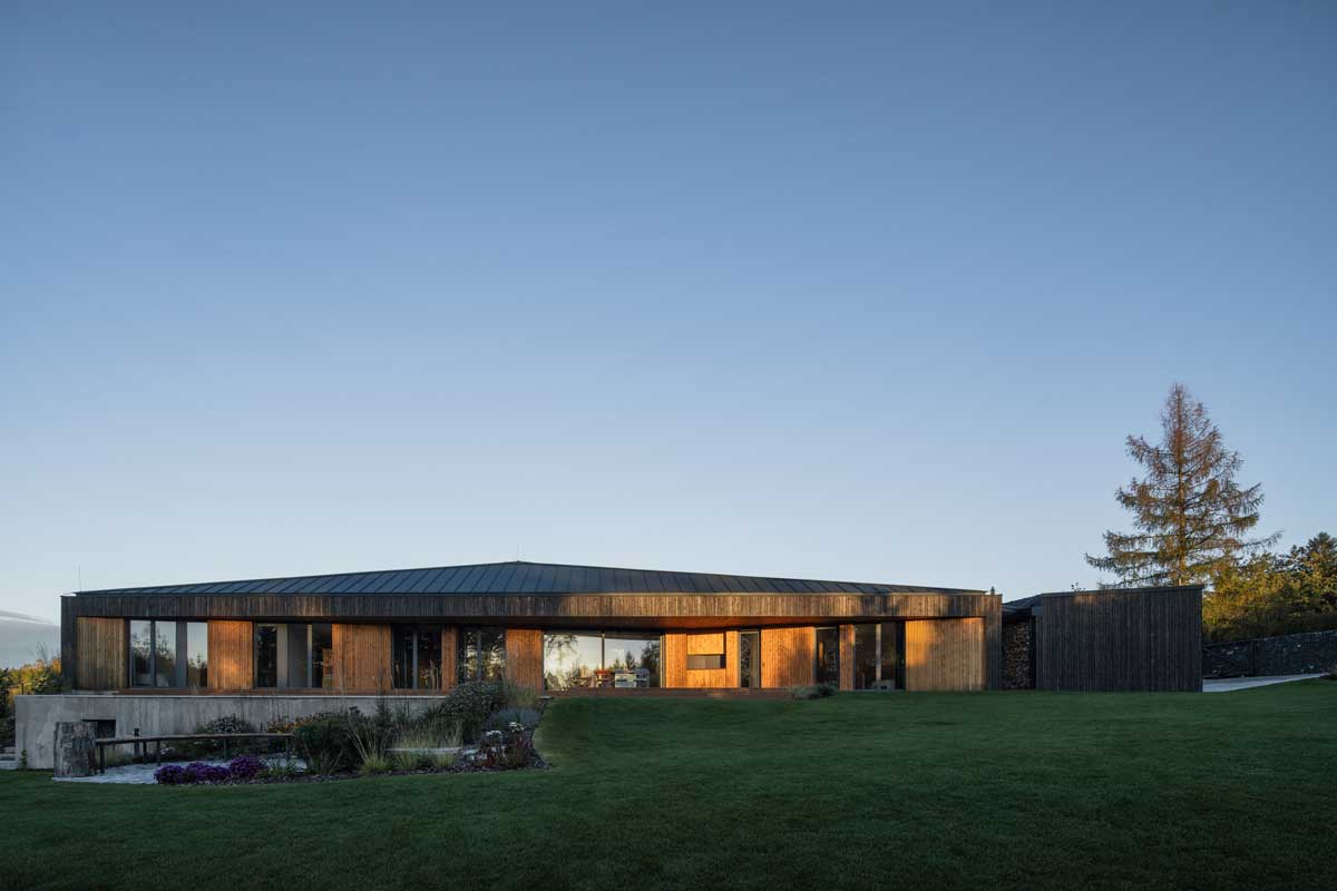 Moderní rodinný dům na Vysočině s dřevěnou fasádou, zapuštěný do svažité zahrady při večerním světle.