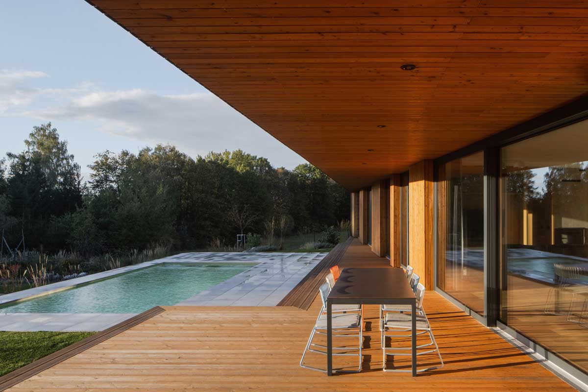 Krytá terasa moderního domu s dřevěnou podlahou, venkovním posezením a zapuštěným bazénem.