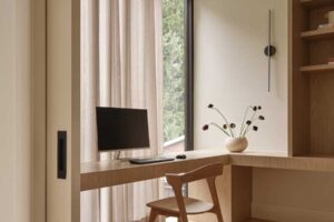 Pracovní kout s dřevěným stolem u okna, otevřenými policemi a minimalistickým osvětlením