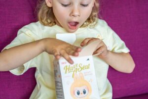 Holčička s nadšeným výrazem otevírá krabičku s figurkou Labubu na pohovce.