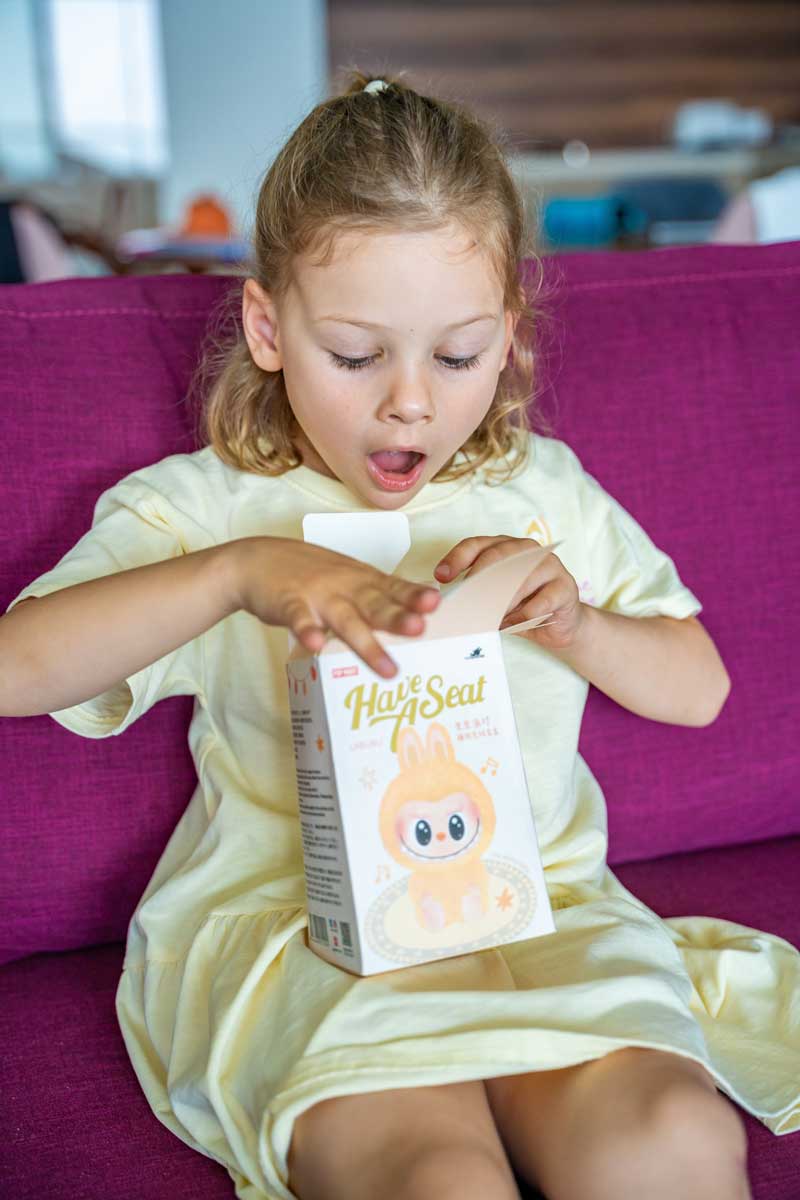 Holčička s nadšeným výrazem otevírá krabičku s figurkou Labubu na pohovce.