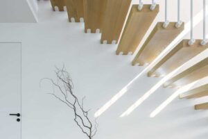 Detail spodní části schodiště s dekorativní vázou a světelnými paprsky dopadajícími z horního patra.