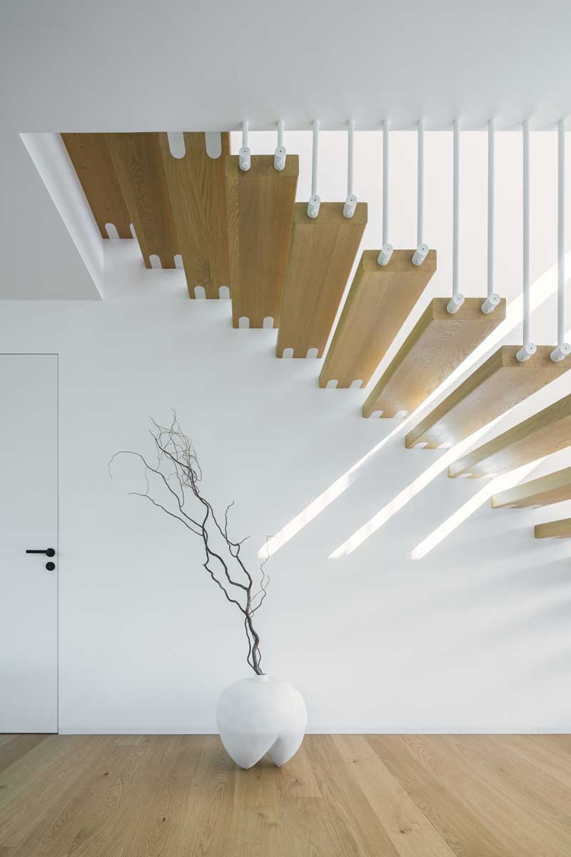 Detail spodní části schodiště s dekorativní vázou a světelnými paprsky dopadajícími z horního patra.