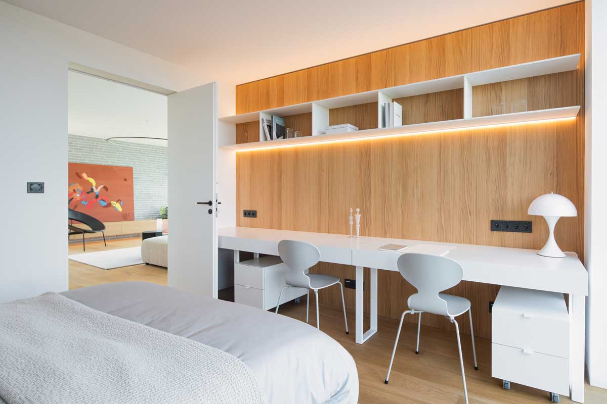 Minimalistický pracovní kout s integrovaným dřevěným obložením a dlouhým bílým stolem pro dvě osoby.