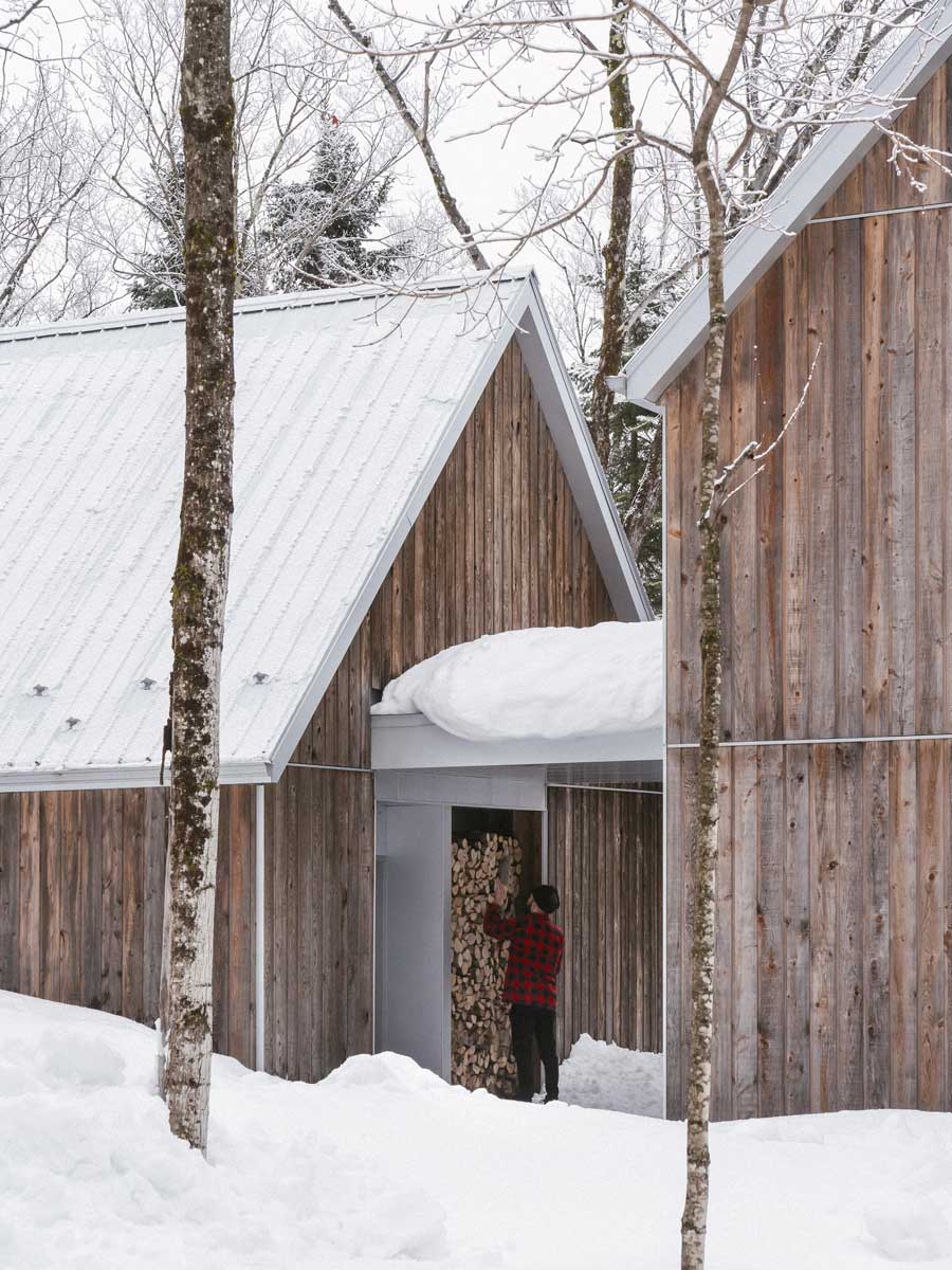 Majitel ukládá dřevo v krytém průchodu mezi dřevěnými objekty domu který přirozeně splývá se zasněženým prostředím