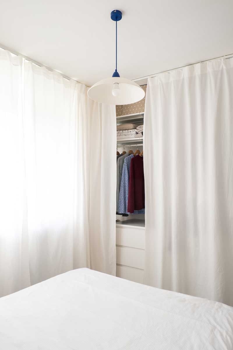 Vestavěné úložné prostory skryté za bílým závěsem v malé ložnici.