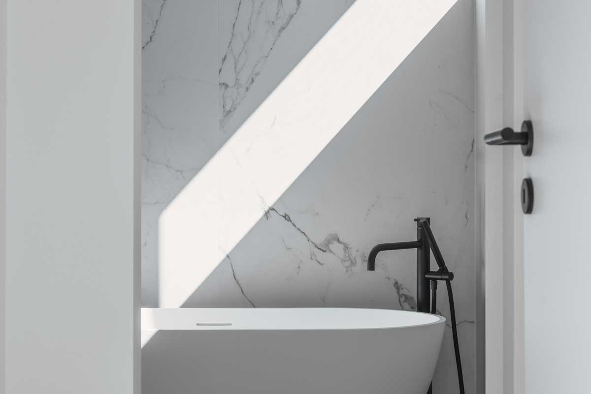 Detail samostatně stojící vany u mramorové stěny s dopadajícím ostrým světlem.