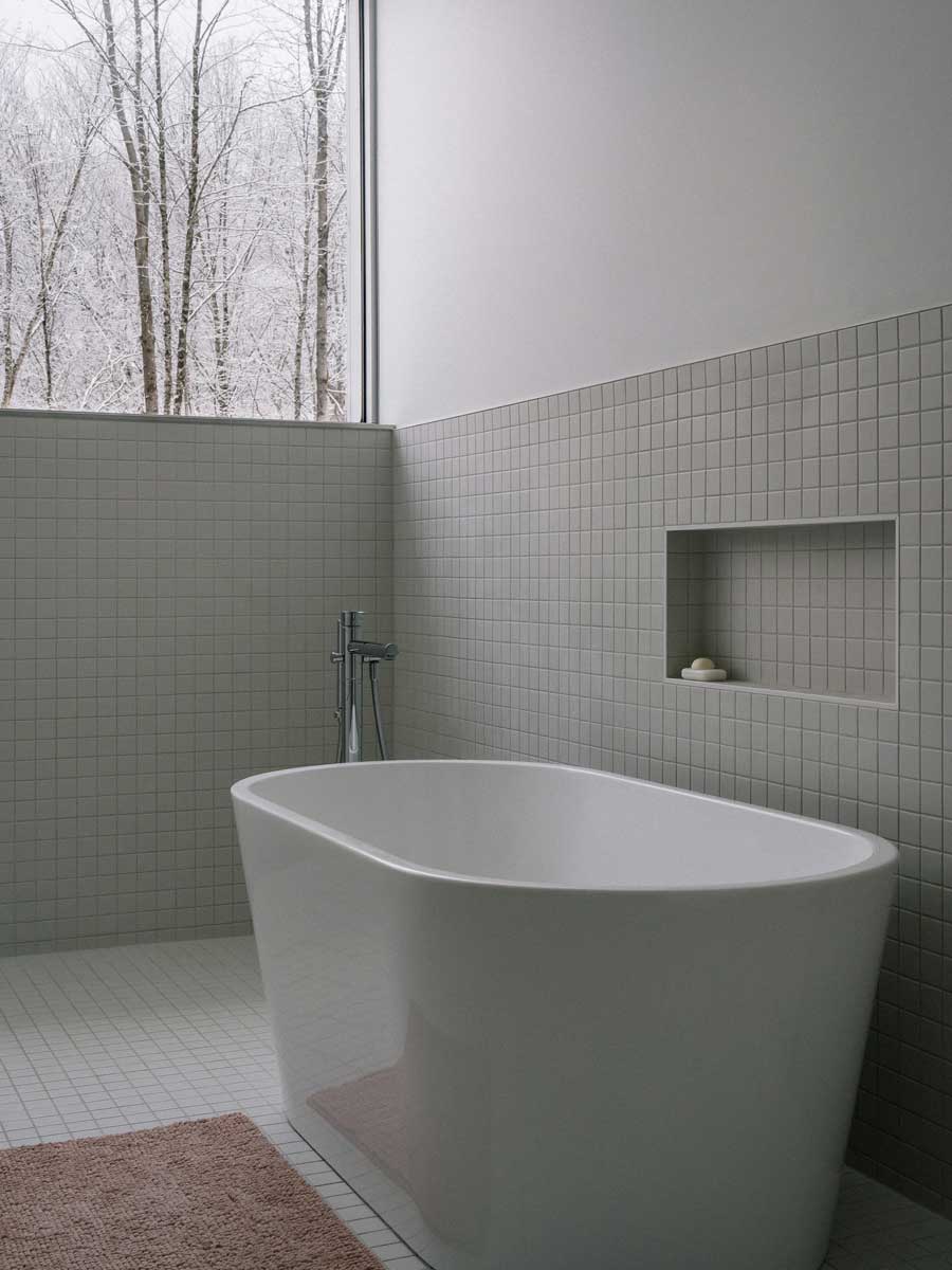 Volně stojící vana v minimalistické koupelně působí jako elegantní vyhlídka do zasněženého lesa a zvýrazňuje propojení interiéru s přírodou