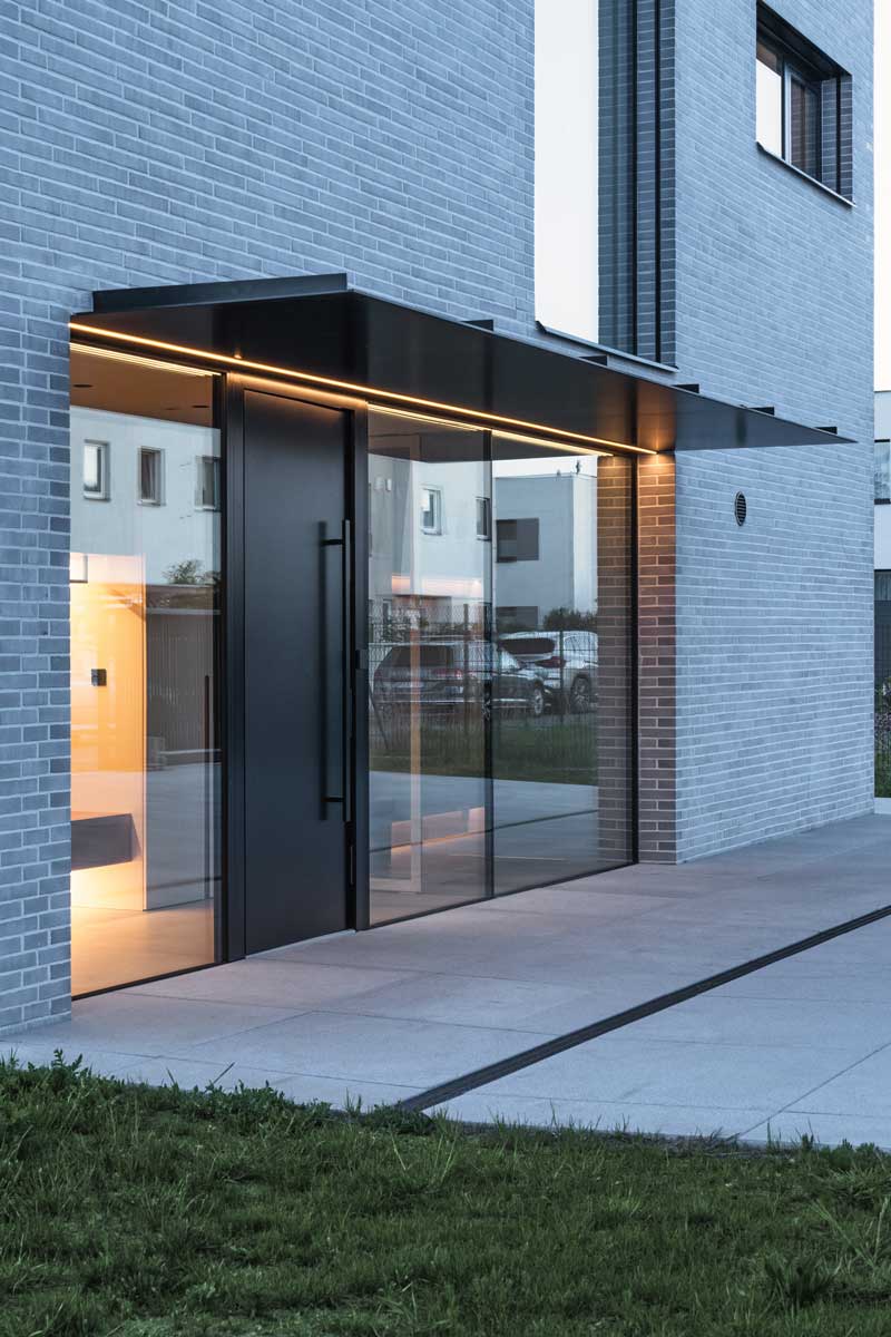 Hlavní vstupní portál domu s prosklenými plochami a černým přístřeškem.