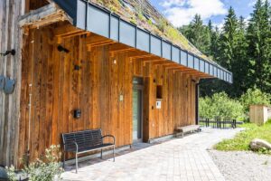 Vstup do horské chaty s dřevěnou fasádou, zelenou střechou a lavičkou pod přesahem střechy