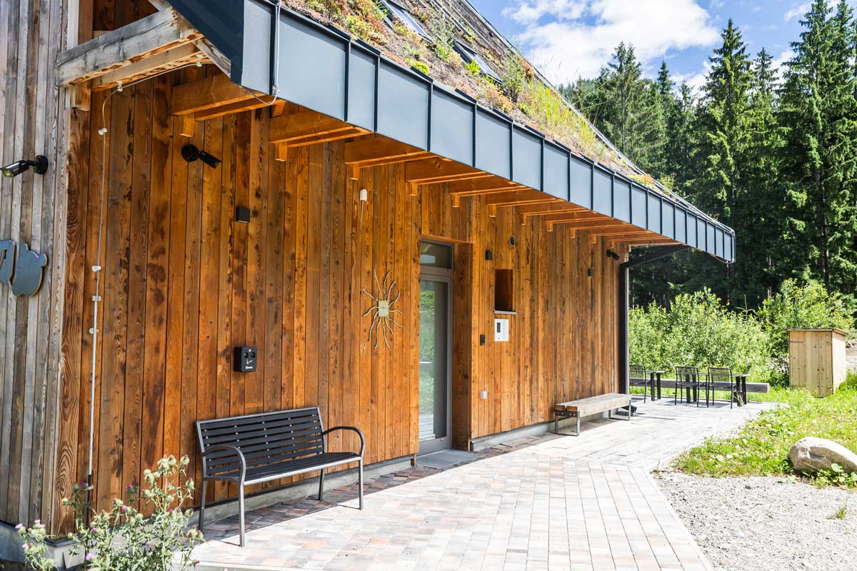 Vstup do horské chaty s dřevěnou fasádou, zelenou střechou a lavičkou pod přesahem střechy