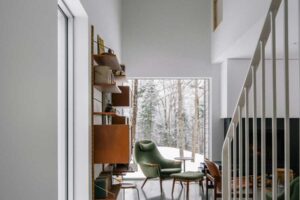 Světlý prostor u schodiště vede do čtecího koutu s ikonickým křeslem a velkým oknem které rámuje zimní les
