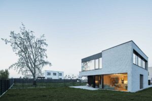 Moderní dvoupodlažní dům s velkými prosklenými plochami otevřený do zahrady při podvečerním světle.