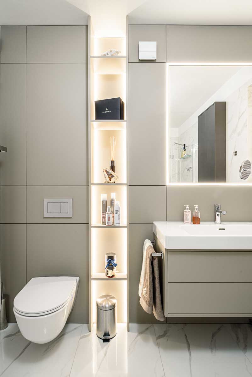 Koupelna s WC, podsvícenými policemi, závěsnou toaletou a umyvadlovou skříňkou v šedém odstínu.
