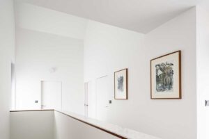 Světlá chodba v horním patře s bílými stěnami, dřevěnými detaily a barevnými uměleckými obrazy.