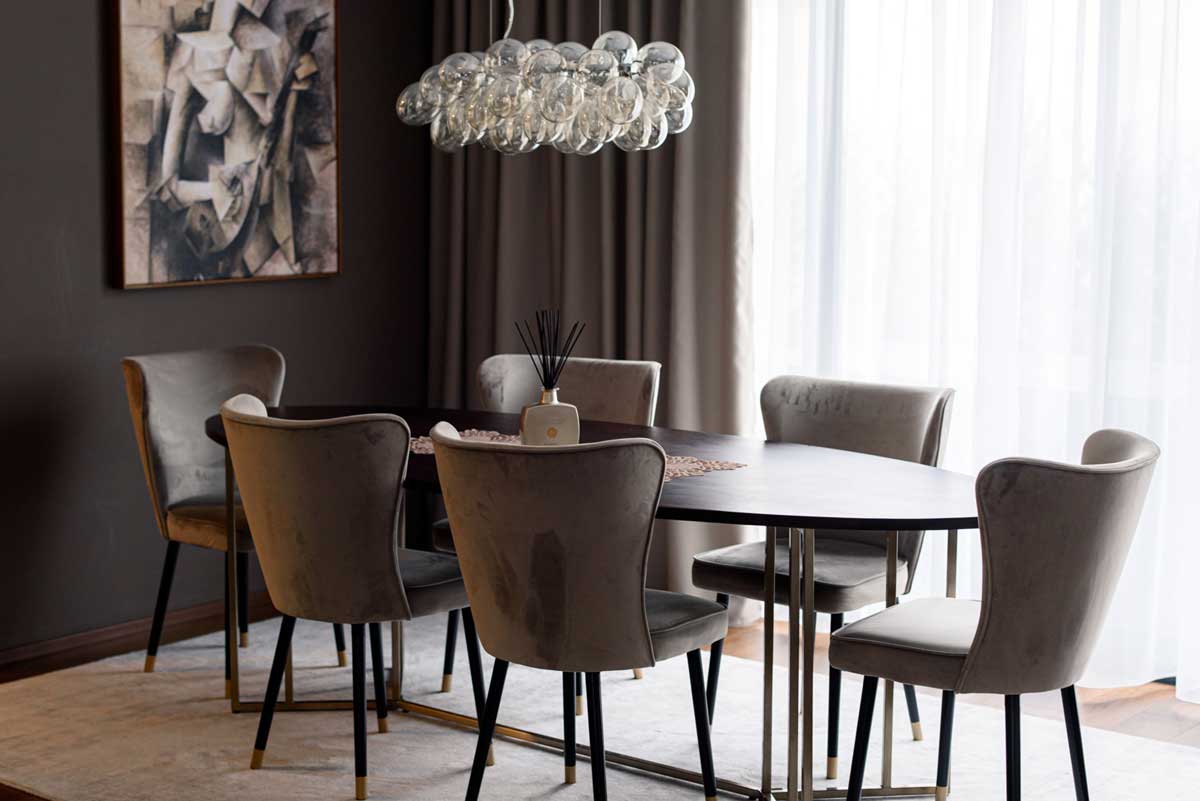 Jídelní kout s oválným stolem z tmavého dřeva, čalouněnými židlemi v šedém odstínu a výrazným skleněným lustrem v minimalistickém interiéru.