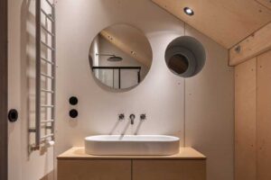 Koupelna v podkroví s dřevěným nábytkem kruhovým zrcadlem a vzorovanou dlažbou