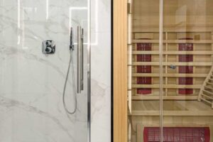 Koupelna se sprchovým koutem a vestavěnou saunou, obložená světlým mramorovým dekorem a dřevem v přírodním odstínu.