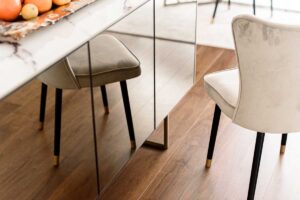 Detail zrcadlových dvířek komody odrážejících podlahu z tmavého dřeva a čalouněné židle.