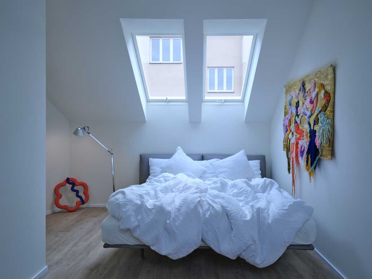 Podkrovní ložnice v pavlačovém domě se střešními okny a důrazem na přirozené denní světlo