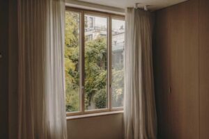 Okno v ložnici s lehkými závěsy a výhledem do zeleně v klidném městském prostředí