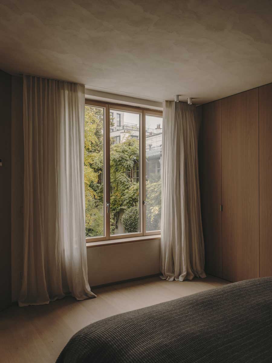 Okno v ložnici s lehkými závěsy a výhledem do zeleně v klidném městském prostředí