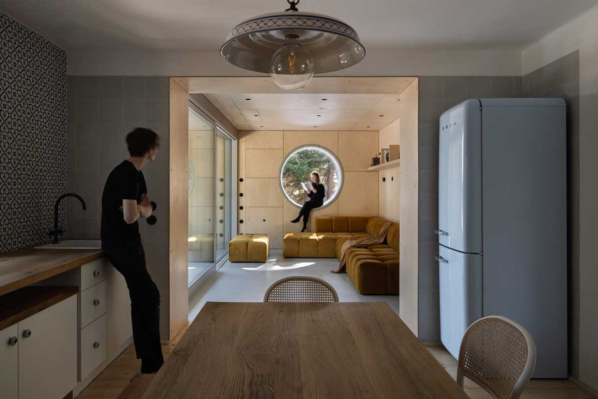 Detail obývacího pokoje s okrovou sedačkou a kruhovým oknem ve dřevěné stěně